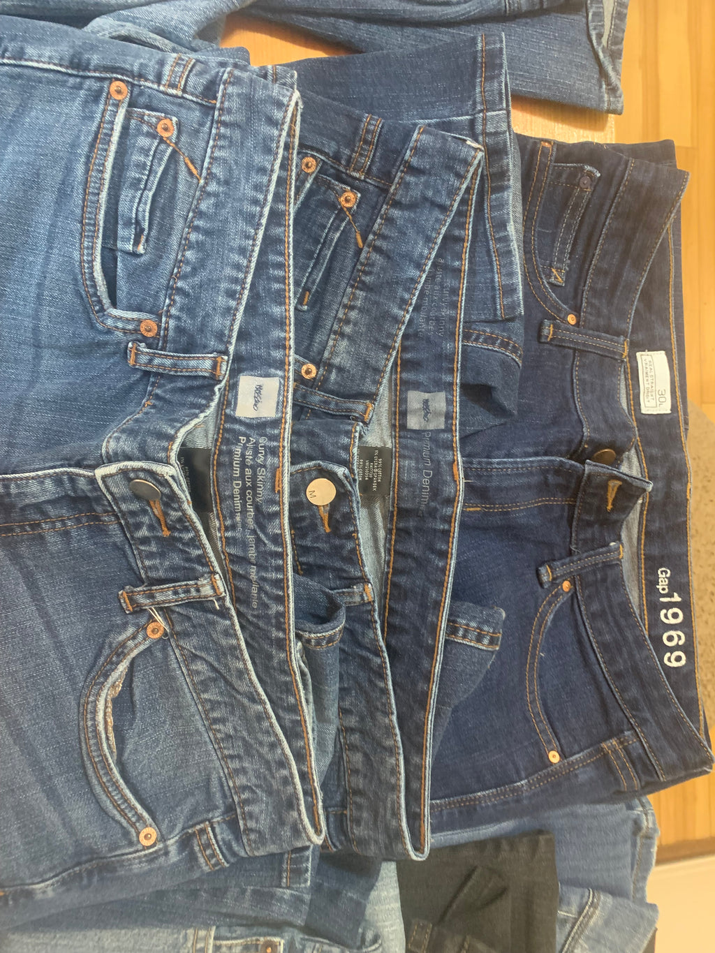 A stack of five blue denim jeans laid flat on a light wood surface, showing various washes from light to dark blue and the waistbands of several brands