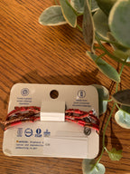 A set of three braided bracelets in red, brown, and black with silver-toned charms, attached to a white cardboard backing card. The card is placed on a wooden table with green eucalyptus leaves in the background.