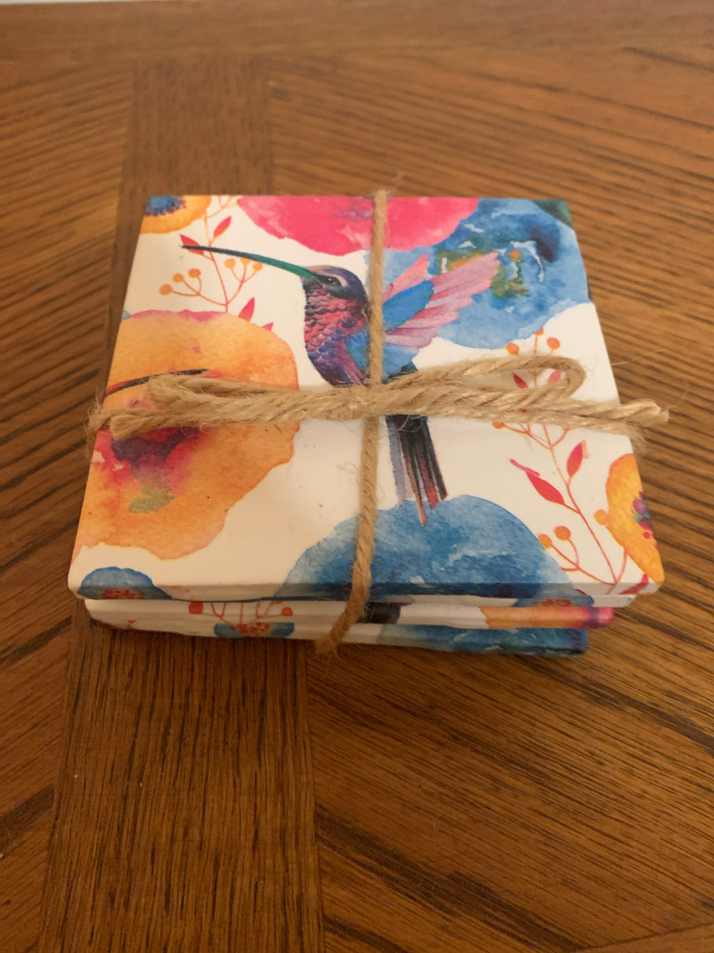A stack of square coasters wrapped in a colorful watercolor design featuring a hummingbird and floral motifs, tied with a rustic jute twine band, placed on a wooden tabletop.