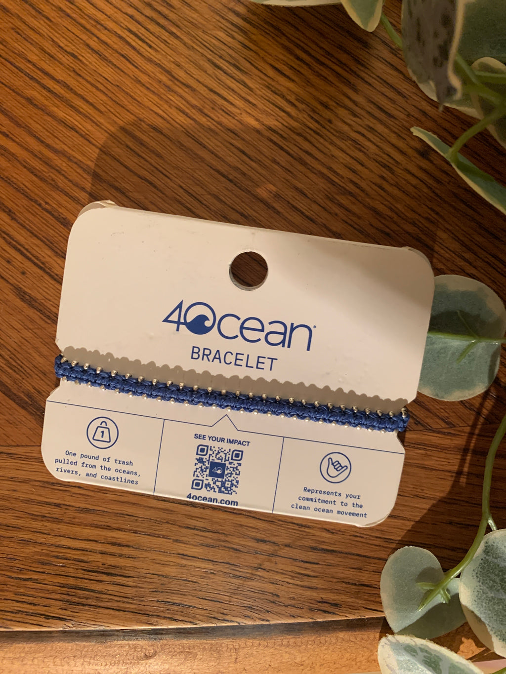 A blue beaded bracelet on a white card labeled '4Ocean Bracelet,' photographed on a wooden surface with green leaves around it.