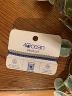 A blue beaded bracelet on a white card labeled '4Ocean Bracelet,' photographed on a wooden surface with green leaves around it.