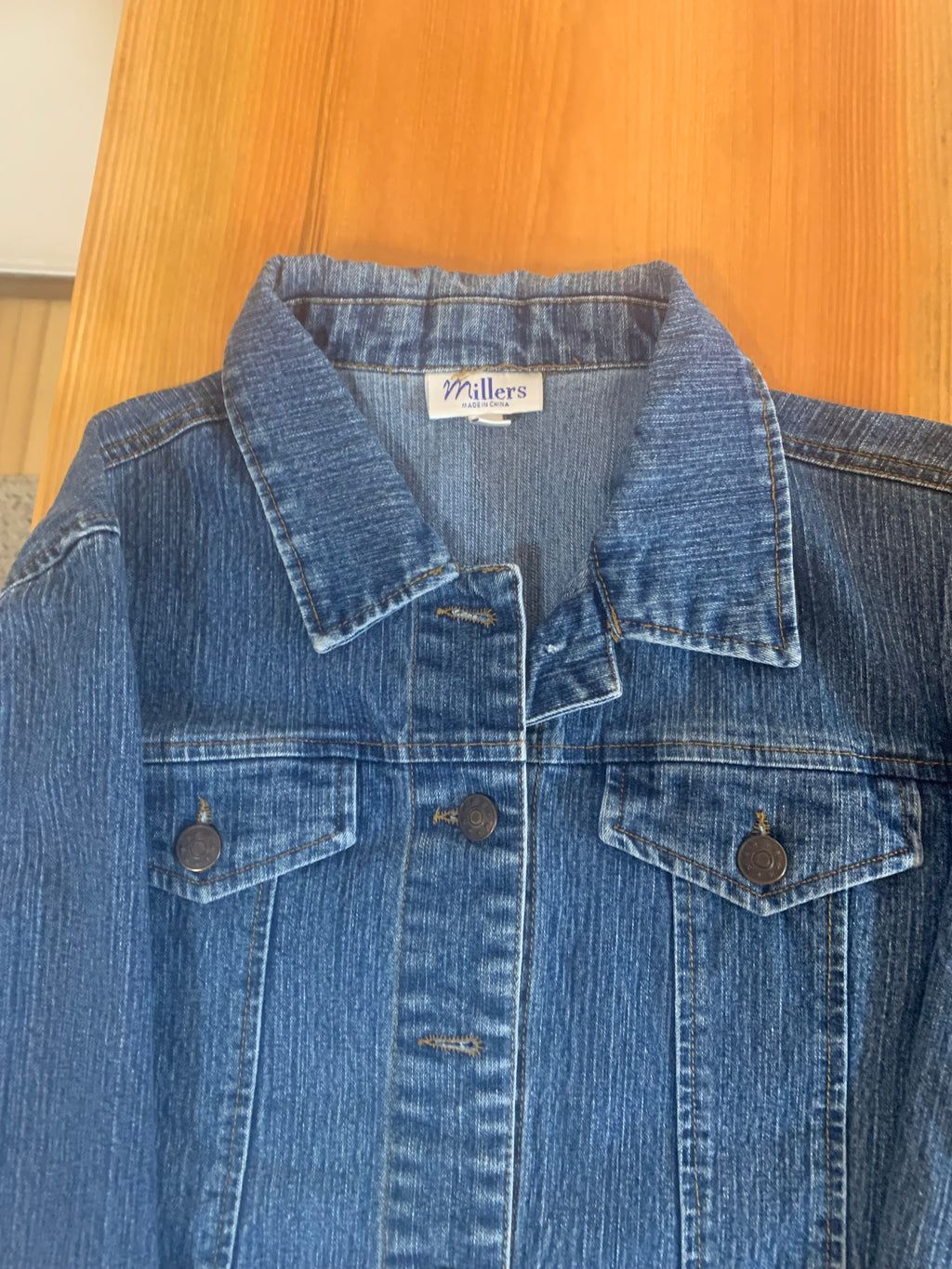 A close-up of a blue denim shirt jacket laid flat on a light wood surface, showing the collar, front button placket, and chest pockets.