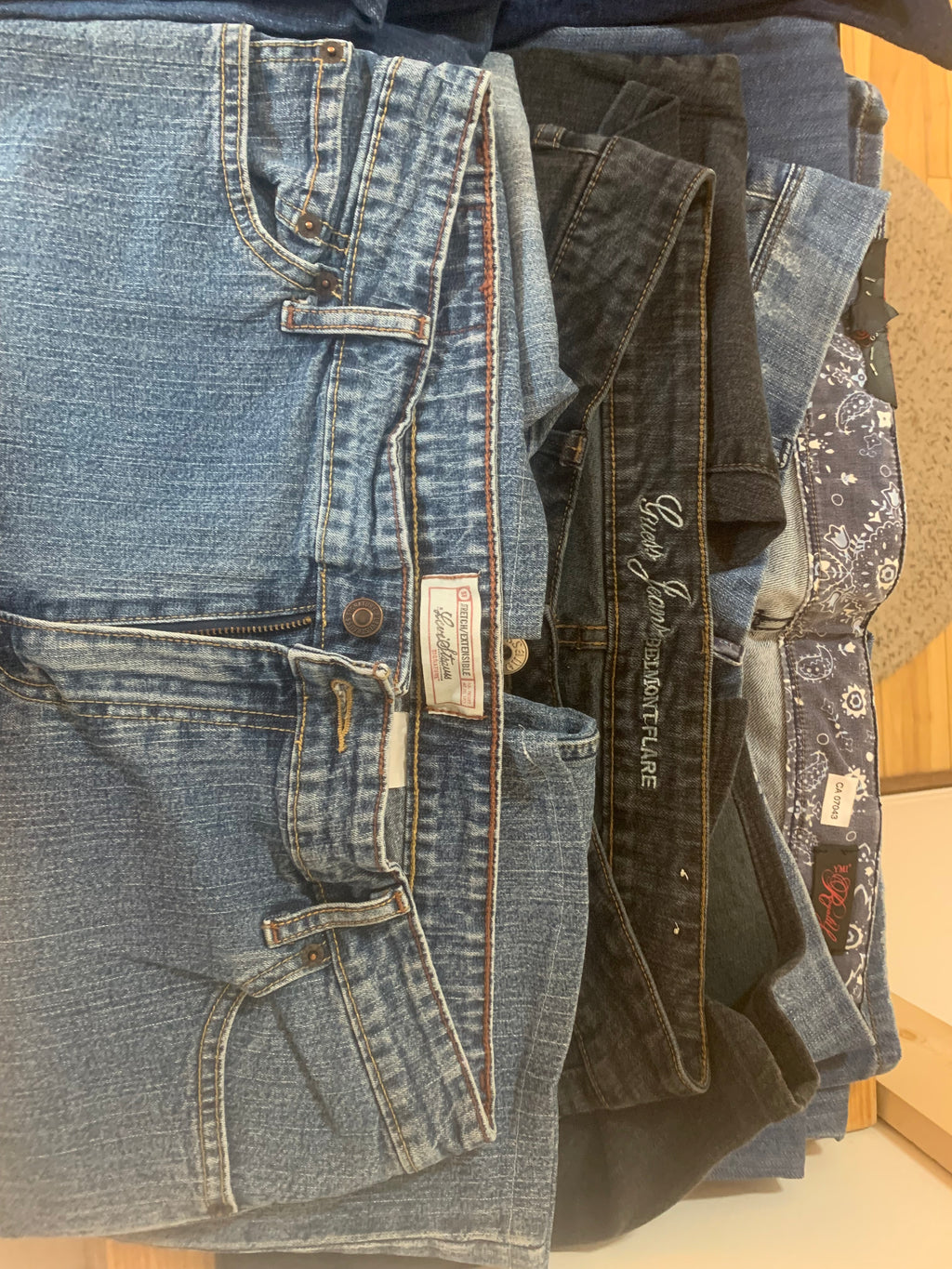 A stack of five blue denim jeans laid flat on a light wood surface, showing various washes from light to dark blue and the waistbands of several brands