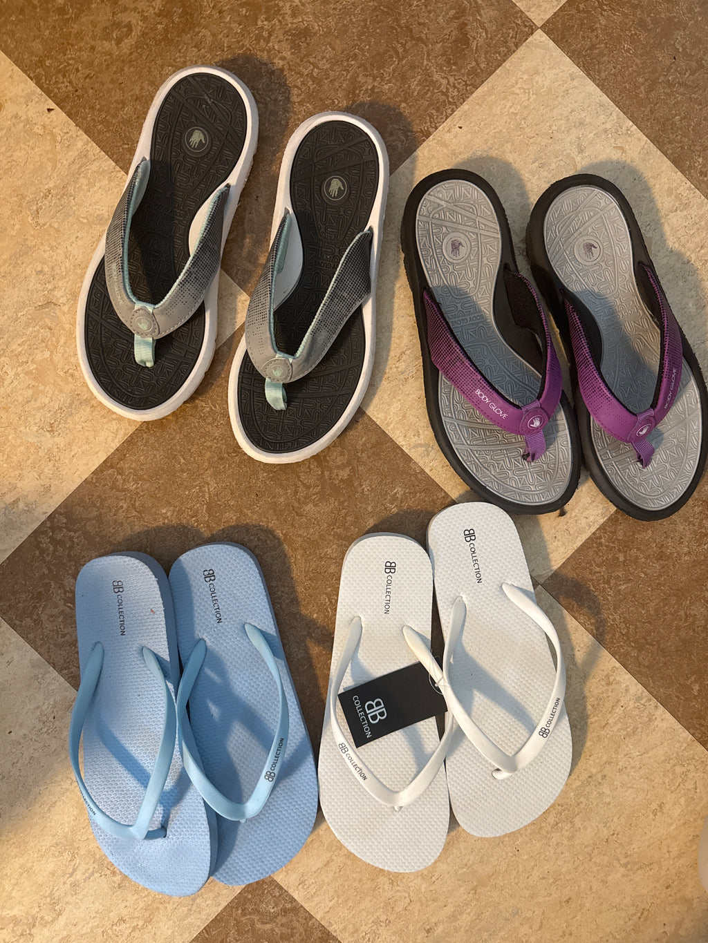 Four pairs of flip-flop sandals laid out on a tan tiled floor: purple and black, white and black, white and black, and light blue and white. 