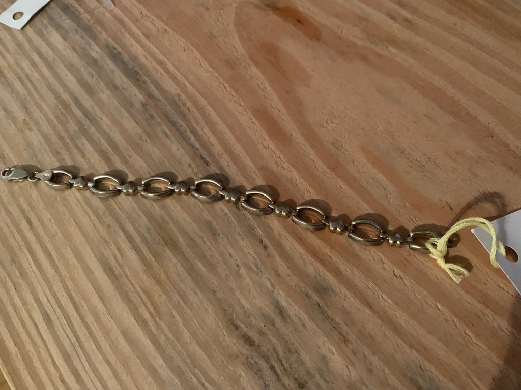 metal chain bracelet with alternating oval and round links, laid diagonally on a wooden surface with a small yellow tag attached.