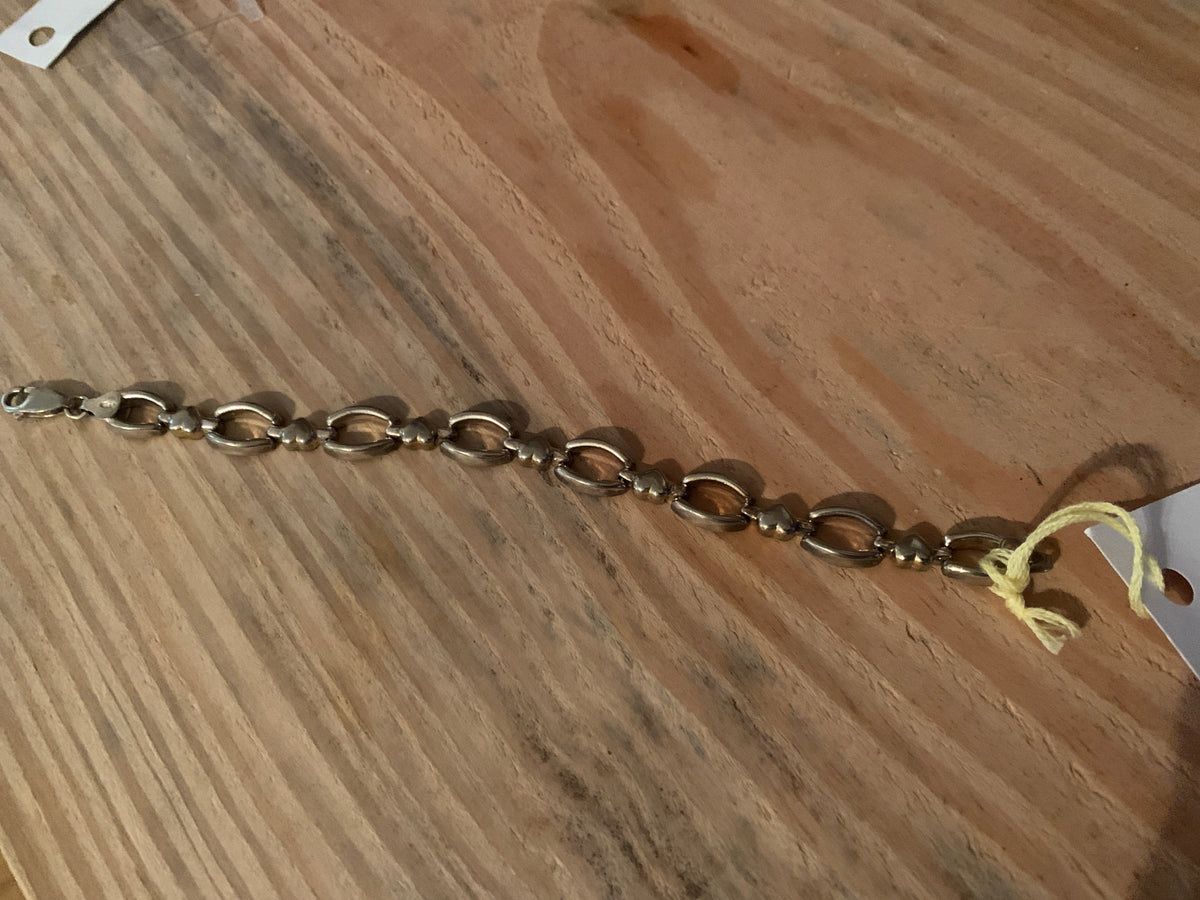 metal chain bracelet with alternating oval and round links, laid diagonally on a wooden surface with a small yellow tag attached.
