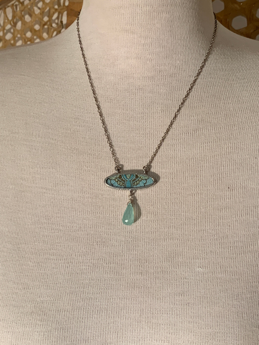 A silver-toned necklace displayed on a cream fabric mannequin bust, featuring an oval pendant with turquoise inlay and a dangling light green teardrop stone.