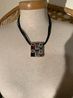 A geometric pendant necklace displayed on a cream fabric mannequin bust. The pendant features a square grid pattern in gray, black, and red, suspended from a black cord.