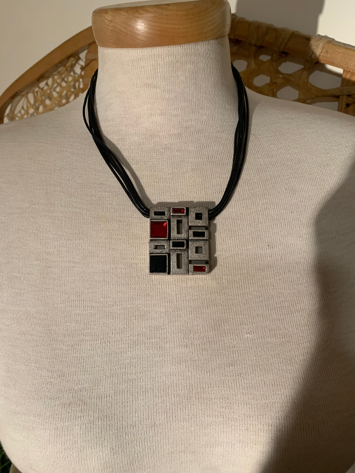 A geometric pendant necklace displayed on a cream fabric mannequin bust. The pendant features a square grid pattern in gray, black, and red, suspended from a black cord.