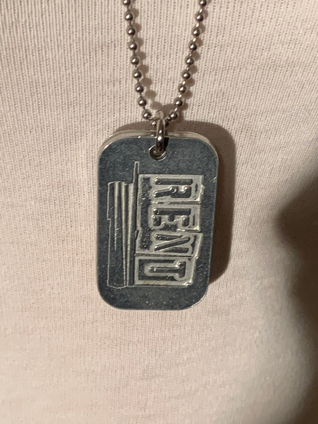 A silver-toned metal dog tag necklace with a ball chain, featuring the word 'REND' in raised block letters on a darkened background, displayed against a light beige fabric backdrop.