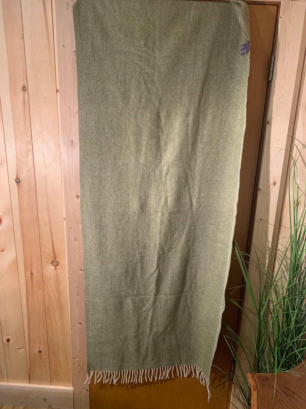 An olive green woven blanket or throw with cream fringe, hanging on a wooden door in a rustic indoor setting with wood paneling and a potted plant.