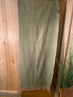 An olive green woven blanket or throw with cream fringe, hanging on a wooden door in a rustic indoor setting with wood paneling and a potted plant.