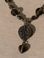 A close-up of a beaded necklace with dark brown and smoky translucent beads, a silver-toned oval medallion pendant, and a dangling heart-shaped bead, laid on a textured beige fabric background.
