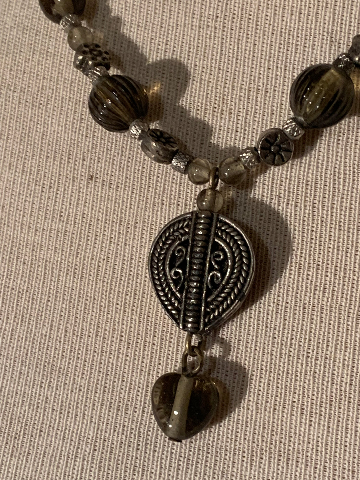 A close-up of a beaded necklace with dark brown and smoky translucent beads, a silver-toned oval medallion pendant, and a dangling heart-shaped bead, laid on a textured beige fabric background.