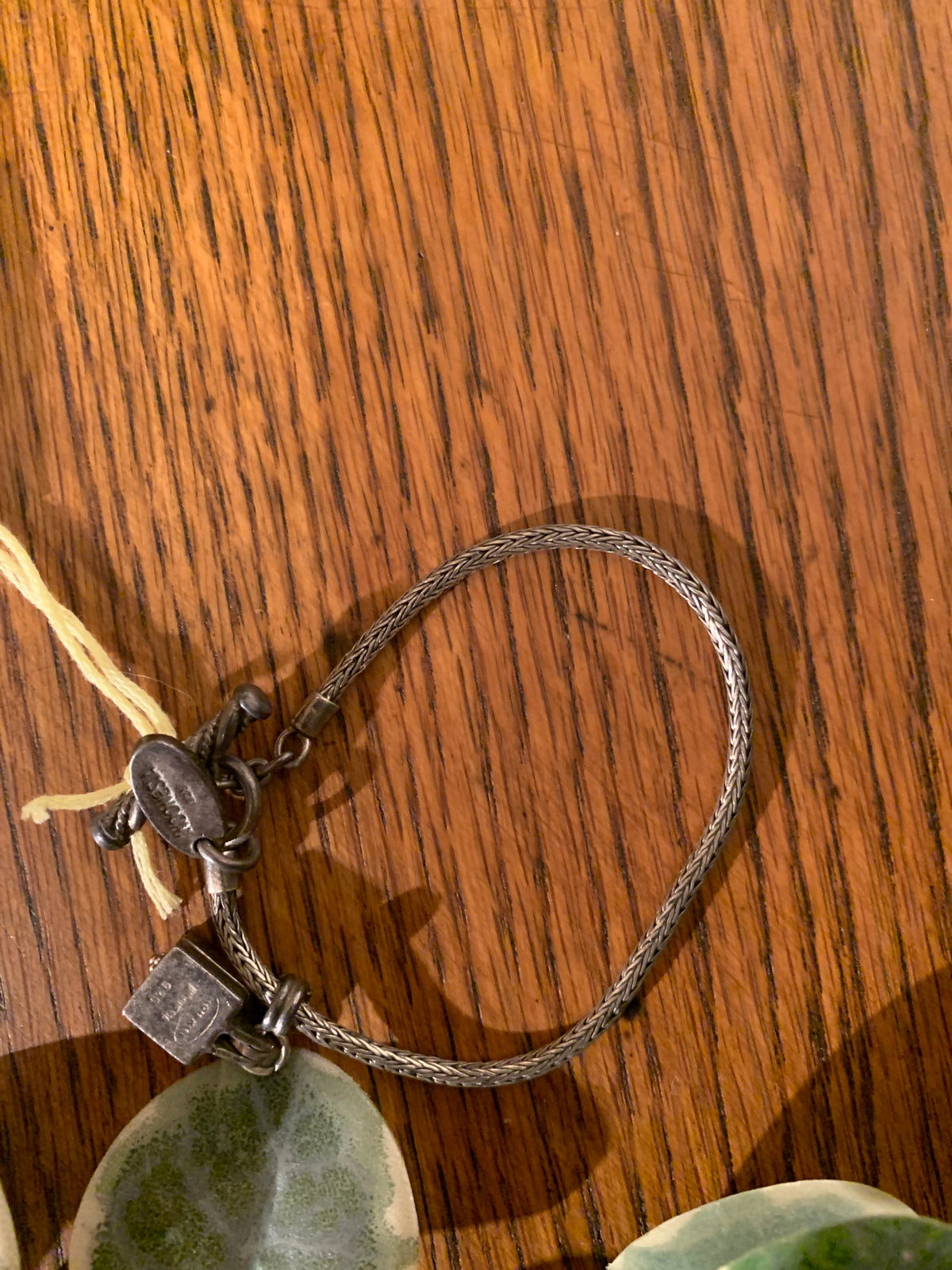 A silver-toned braided cord bracelet with a toggle clasp and small hanging tag, photographed on a warm brown wood surface.