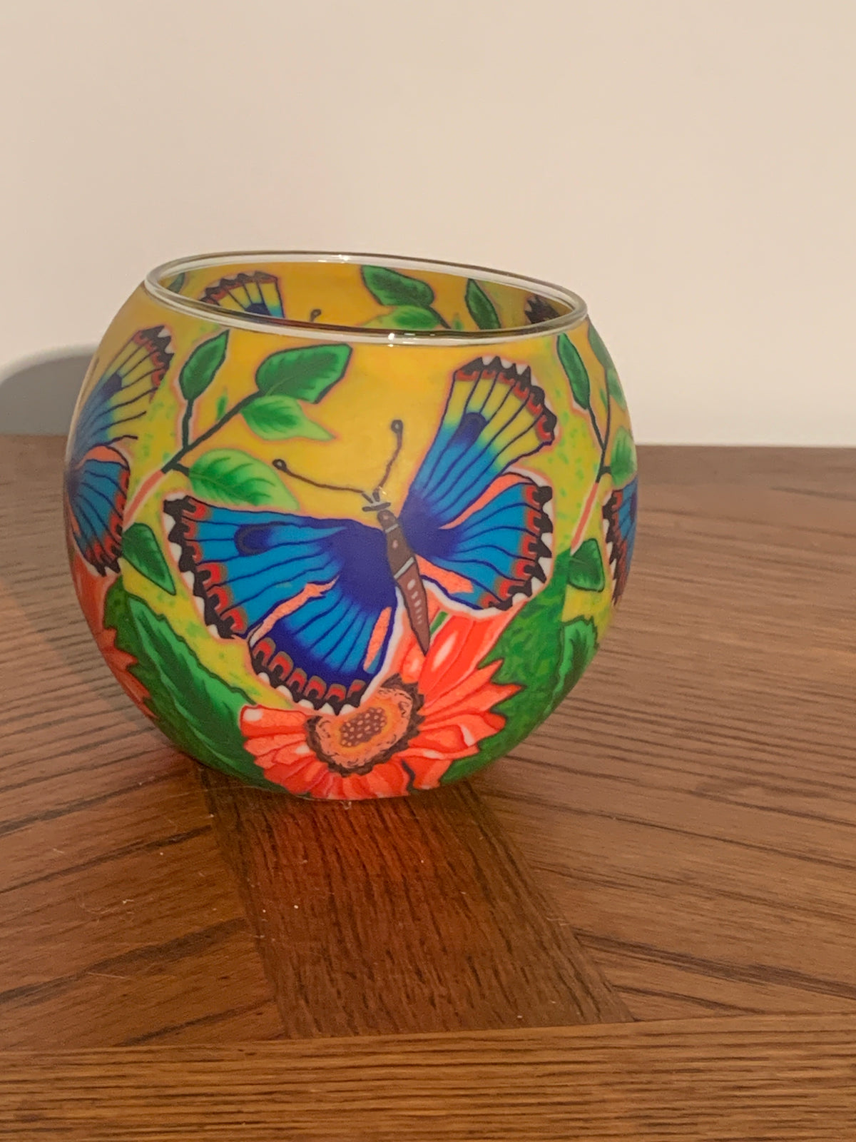 A round glass candle holder with a vibrant hand-painted design featuring a blue and orange butterfly, green leaves, and a red flower on a yellow background, sitting on a wooden table.