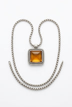 A silver-toned chain necklace with a square pendant featuring a central amber-orange stone, displayed on a textured off-white fabric background.