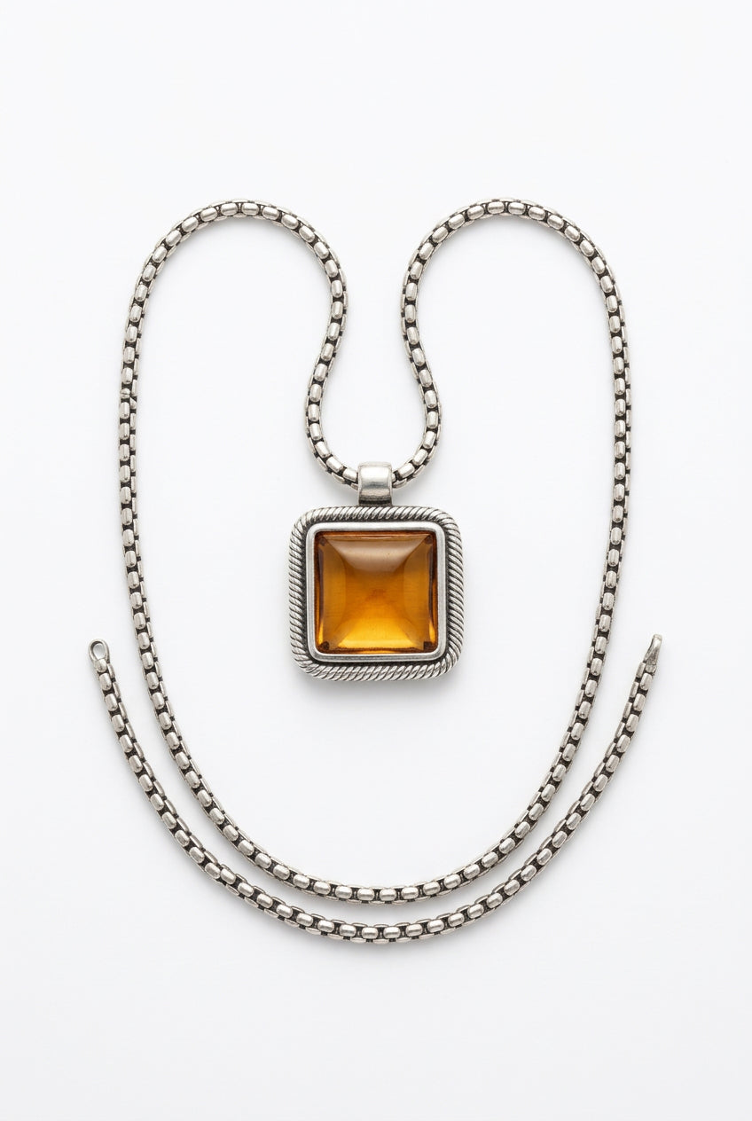 A silver-toned chain necklace with a square pendant featuring a central amber-orange stone, displayed on a textured off-white fabric background.