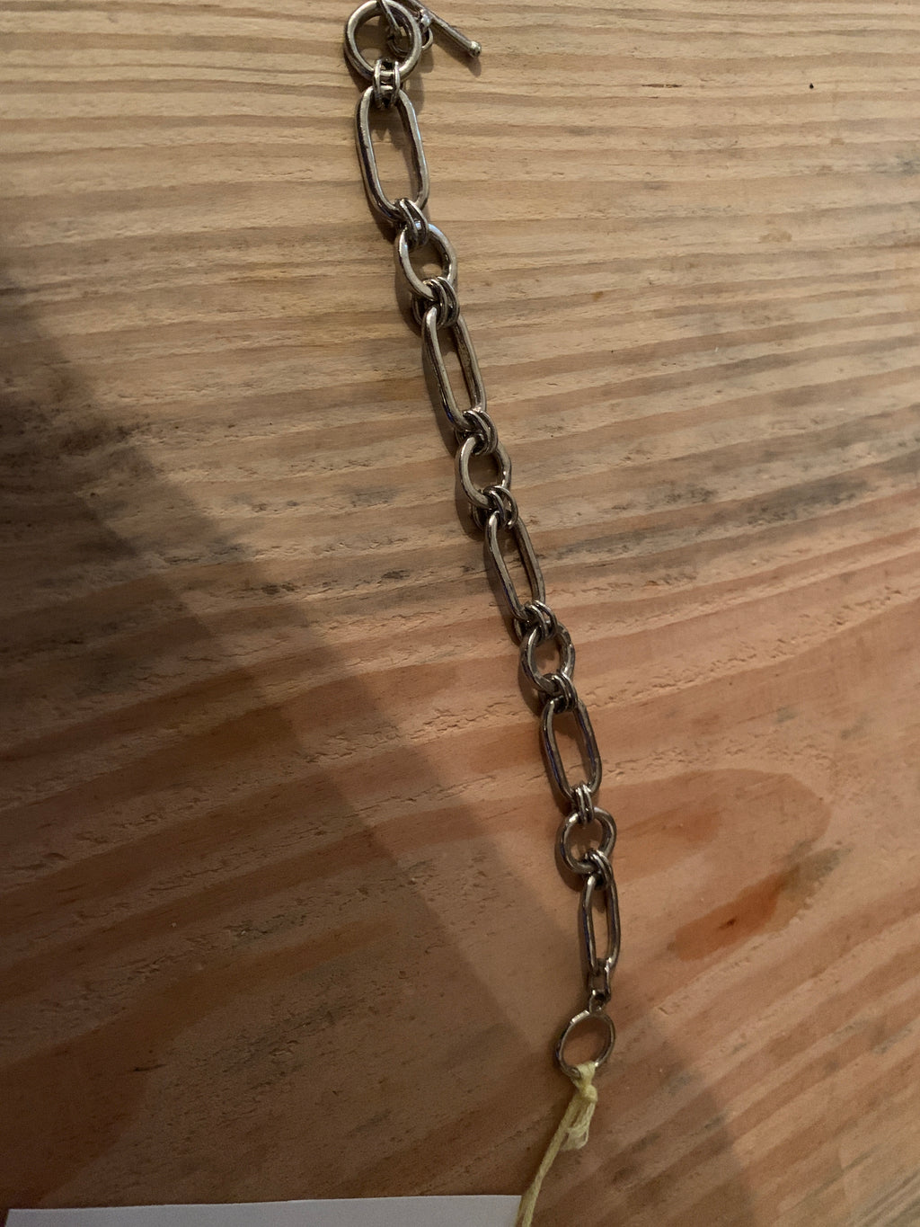 A dark metallic chain link bracelet or anklet laid diagonally on a light brown wooden surface.