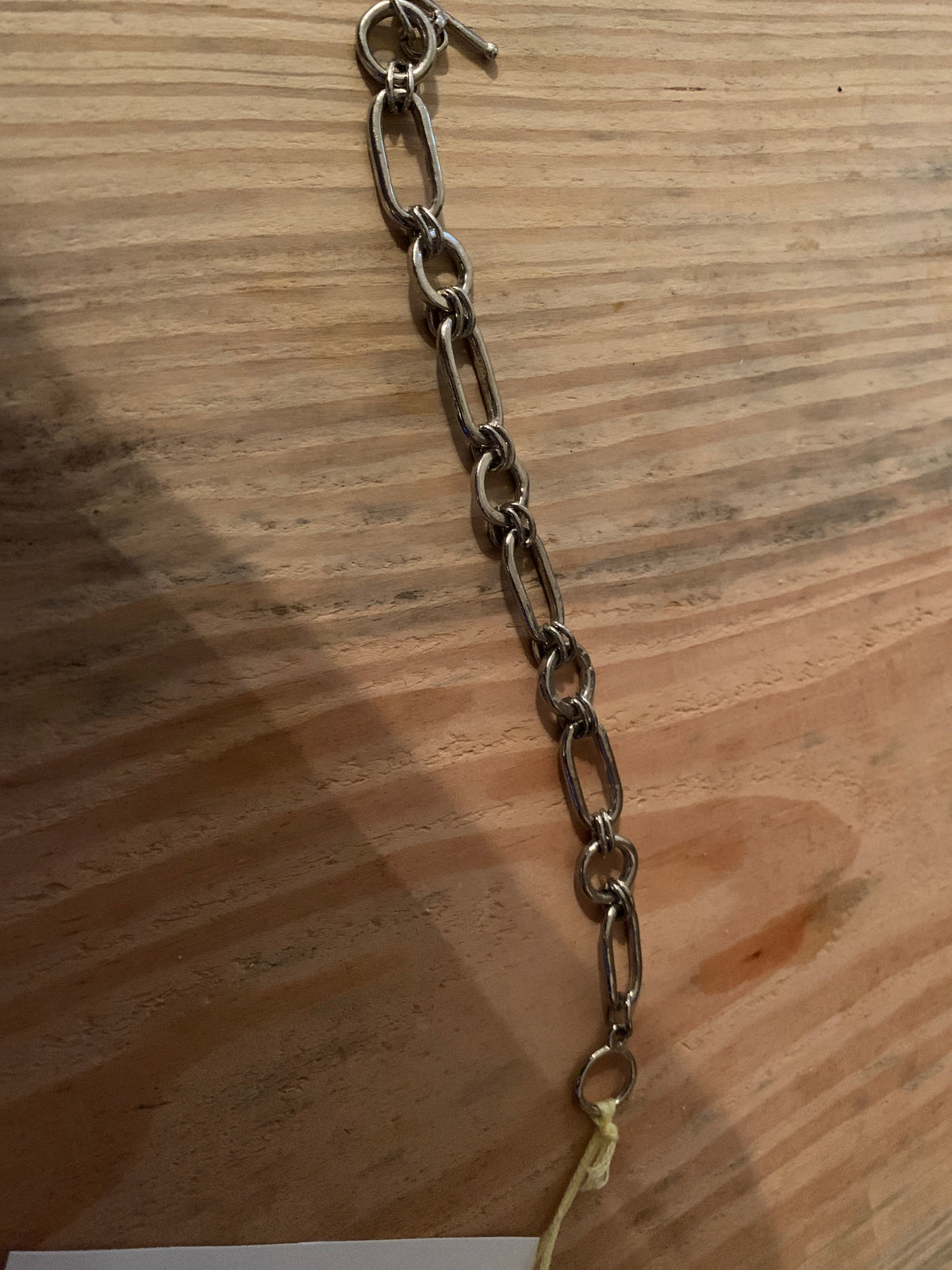 A dark metallic chain link bracelet or anklet laid diagonally on a light brown wooden surface.