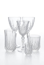 A set of clear cut-crystal and ribbed glass tableware pieces arranged on a wooden tabletop, with a card reading MARQUIS visible behind them and potted greenery in the background.