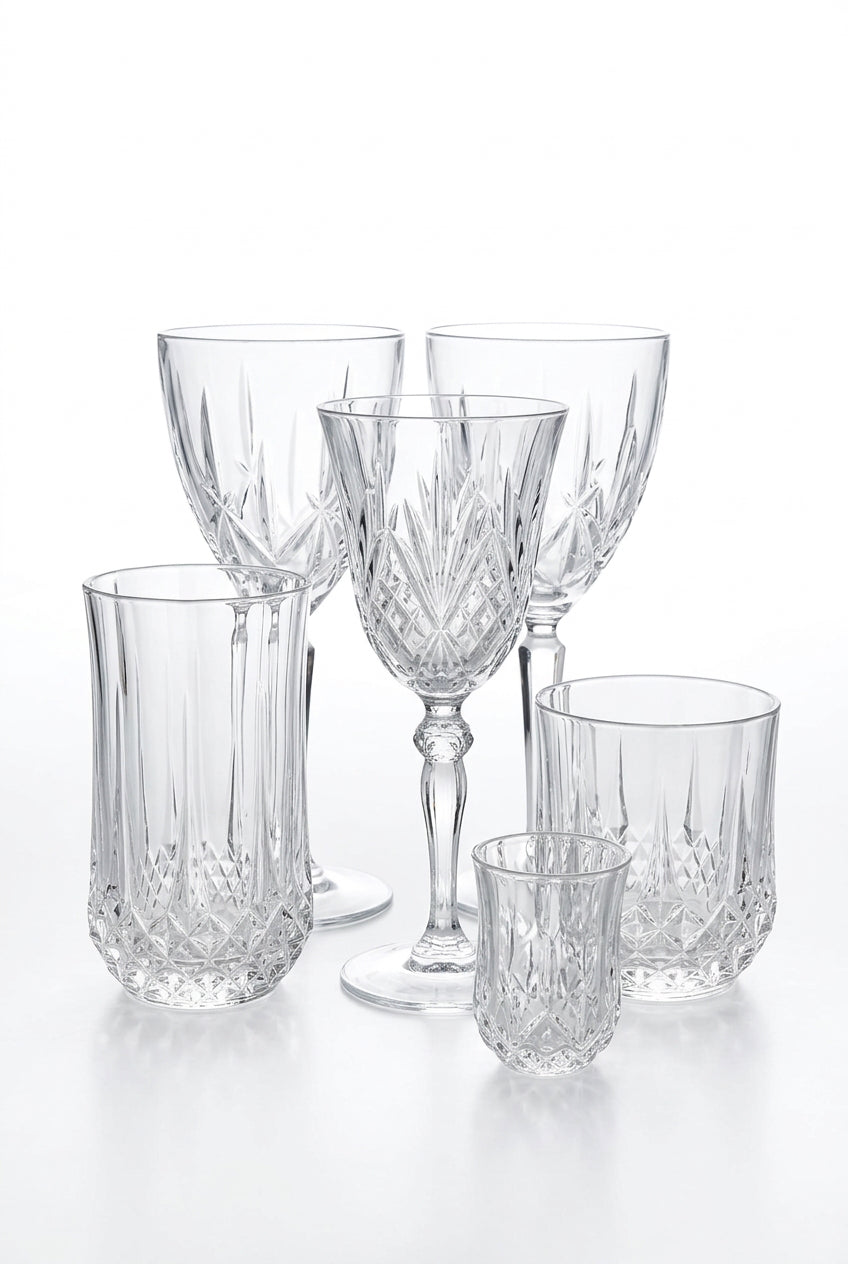 A set of clear cut-crystal and ribbed glass tableware pieces arranged on a wooden tabletop, with a card reading MARQUIS visible behind them and potted greenery in the background.