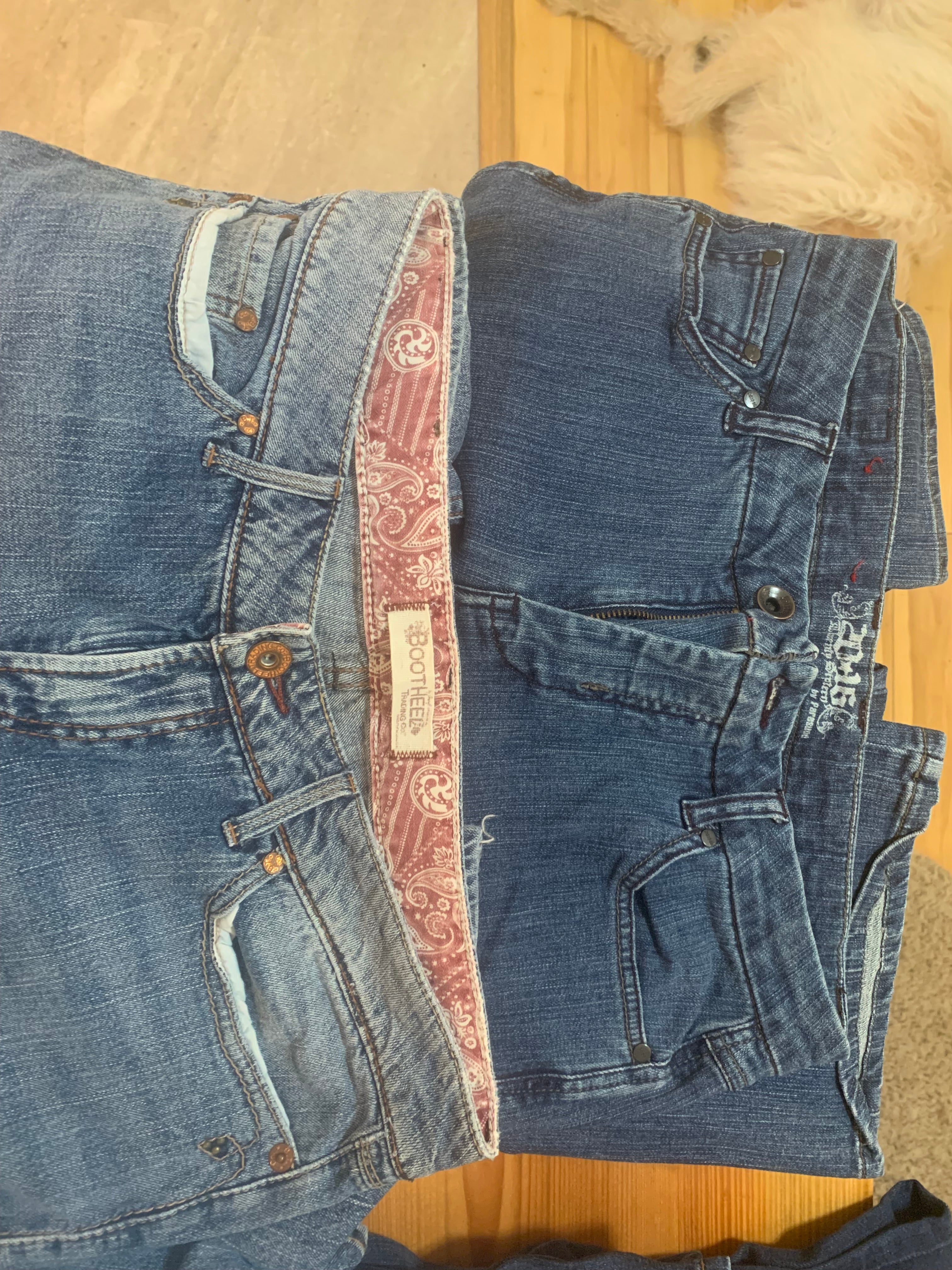Two pairs of folded blue jeans laid flat on a light wood floor. The top pair is a darker medium-wash blue, and the bottom pair is a lighter washed blue denim 