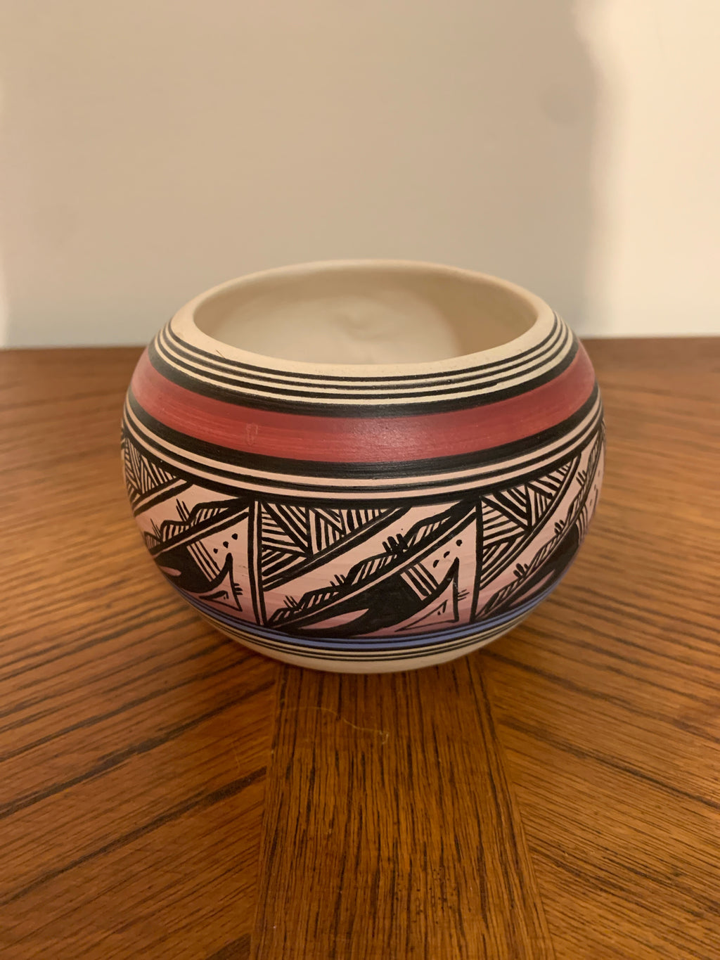 A small round ceramic pot with a Southwestern geometric pattern, featuring black, pink, and red bands on a wooden table against a plain wall.
