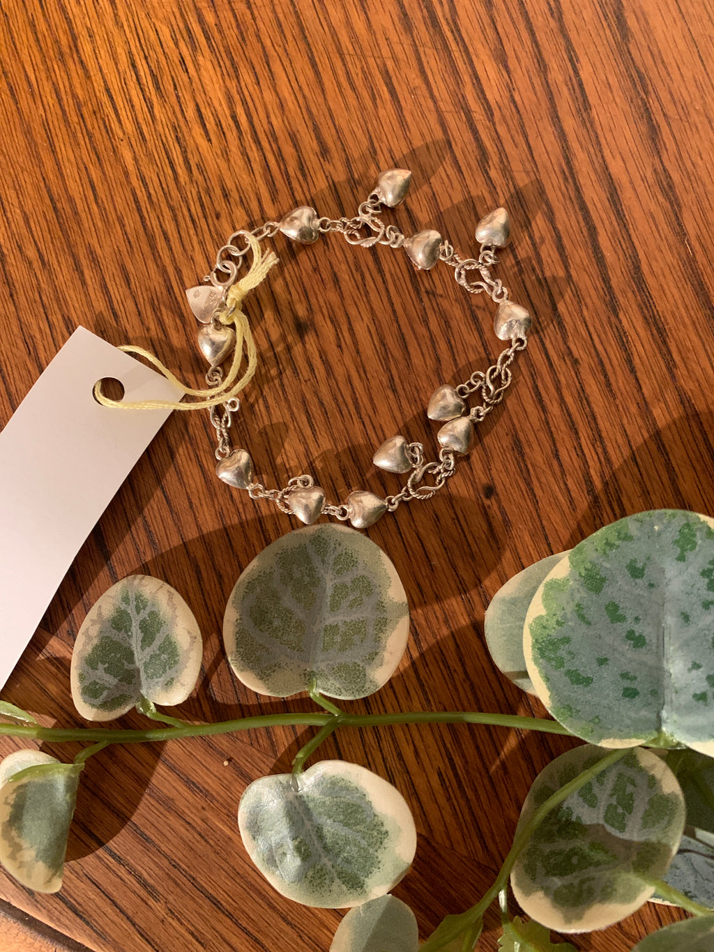 A silver chain bracelet decorated with small dangling heart charms, laid on a wooden surface next to a white paper tag and green variegated eucalyptus leaves.
