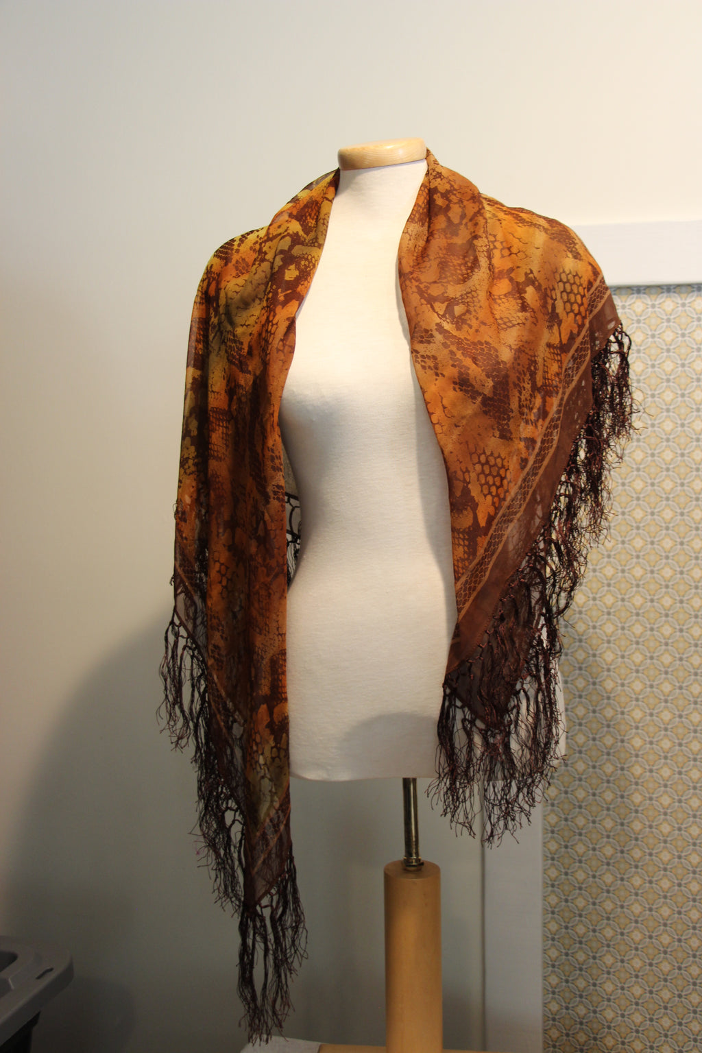 A patterned shawl scarf in shades of brown and orange with green. The scarf features a detailed animal print and long dark fringe along the edges.