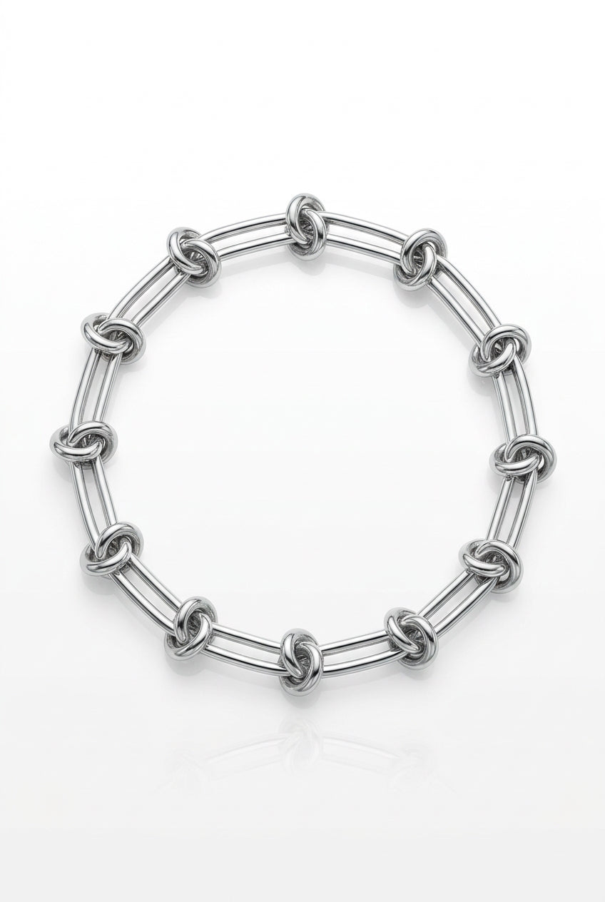 A silver-toned chain link bracelet with elongated oval links, resting on a light wood surface next to a plant and a small paper tag.