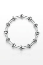 A silver-toned chain link bracelet with elongated oval links, resting on a light wood surface next to a plant and a small paper tag.