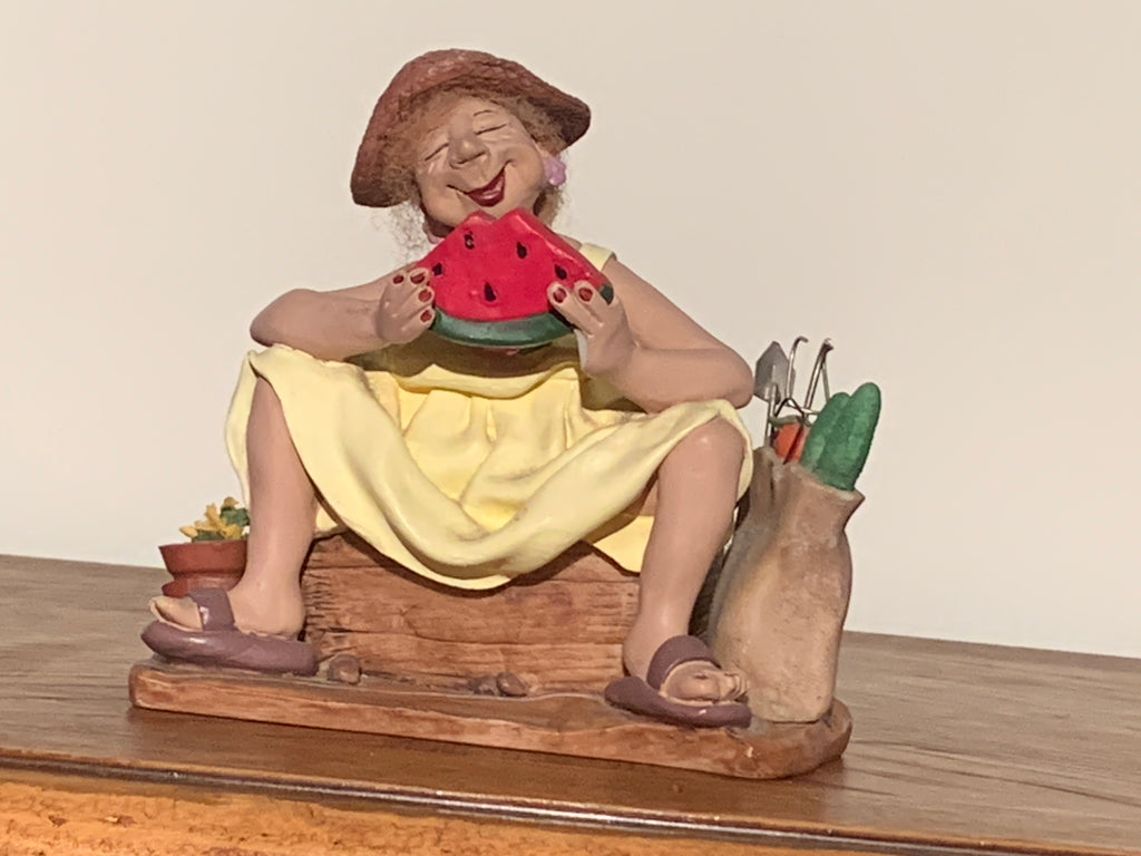 A ceramic garden figurine of a woman sitting on a wooden bench and eating a slice of watermelon, with gardening tools in a pot beside her, displayed on a wooden surface against a plain beige wall.