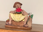 A ceramic garden figurine of a woman sitting on a wooden bench and eating a slice of watermelon, with gardening tools in a pot beside her, displayed on a wooden surface against a plain beige wall.
