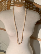A long strand of cream-colored freshwater pearls draped over a beige fabric mannequin torso, set against a rustic wicker chair background.