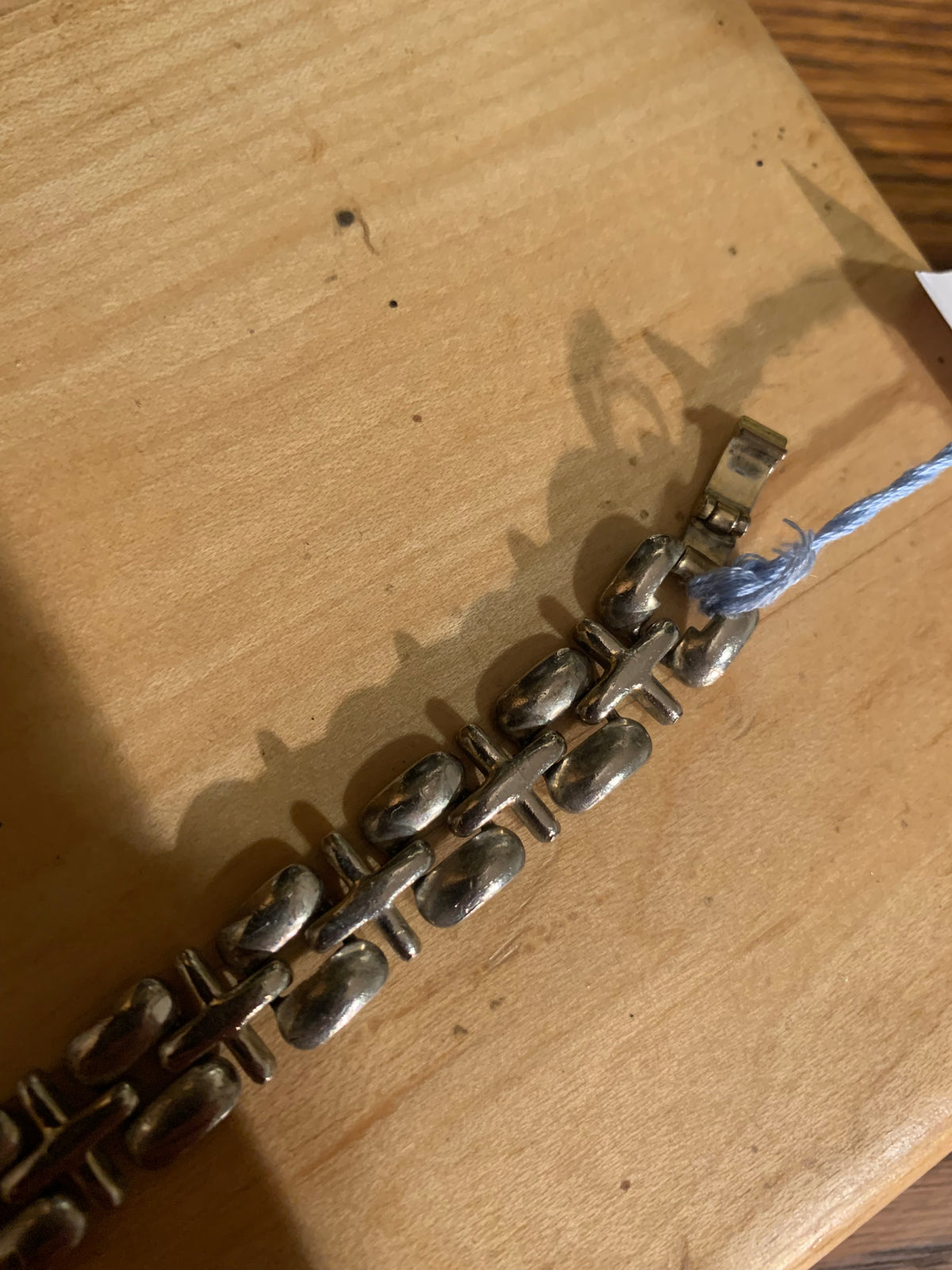 A silver-toned metal bracelet with a unique textured link design, resting on a light wood surface with a blue string tag attached.