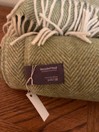 A rolled sage green herringbone wool blanket with white fringe, featuring a dark purple 'Recycled Wool' brand tag on a wooden surface.