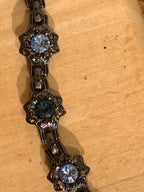 A close-up of a dark metal bracelet with ornate filigree links and round faceted gemstones in light blue, dark teal, and light blue, laid on a light wood surface.