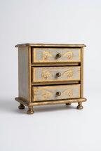 A small vintage-style three-drawer jewelry box or cabinet with a distressed gray-blue painted finish, gold-toned scrollwork accents on the drawer fronts, and dark metal round knobs, shown standing on a wooden surface against a plain light wall.