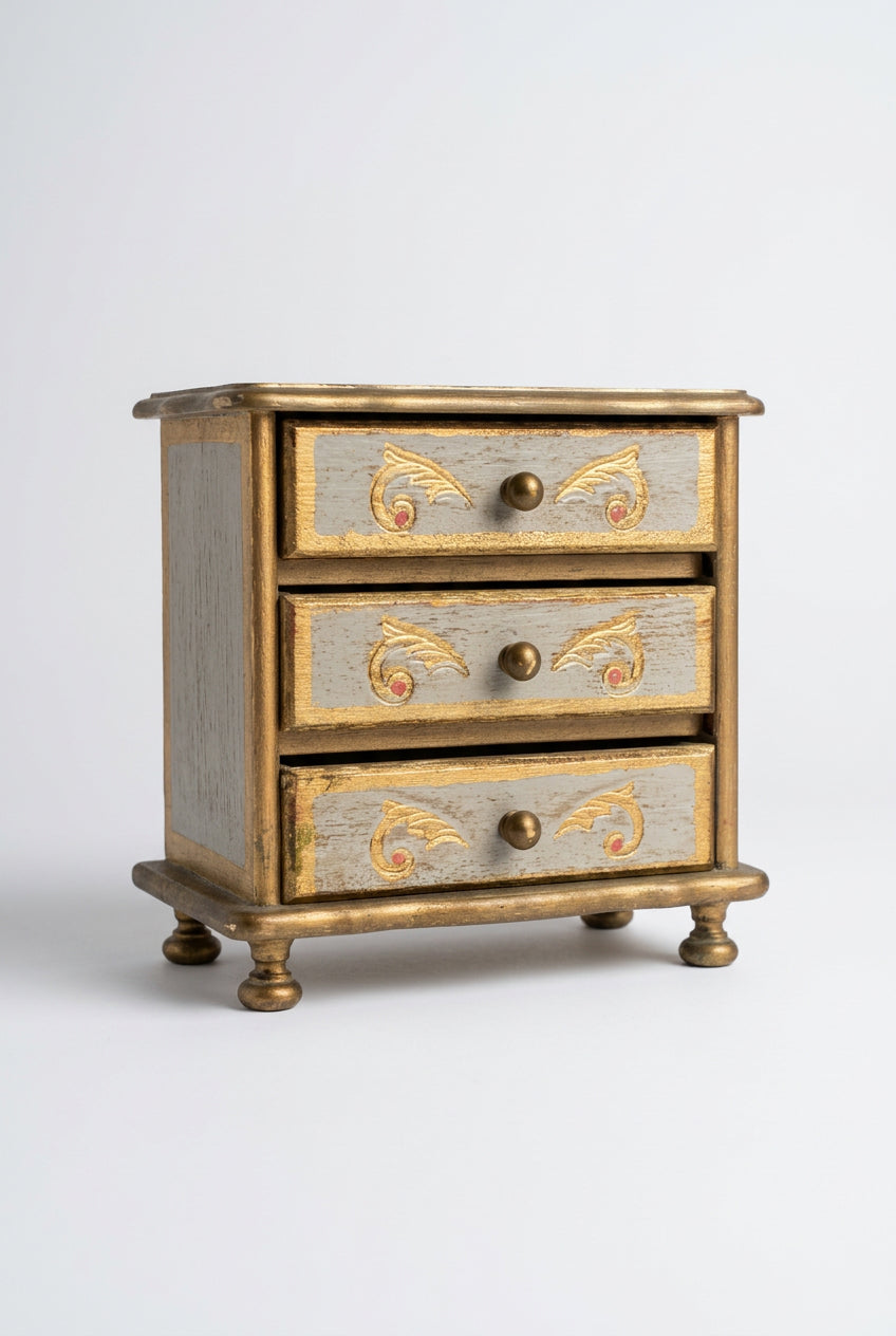 A small vintage-style three-drawer jewelry box or cabinet with a distressed gray-blue painted finish, gold-toned scrollwork accents on the drawer fronts, and dark metal round knobs, shown standing on a wooden surface against a plain light wall.