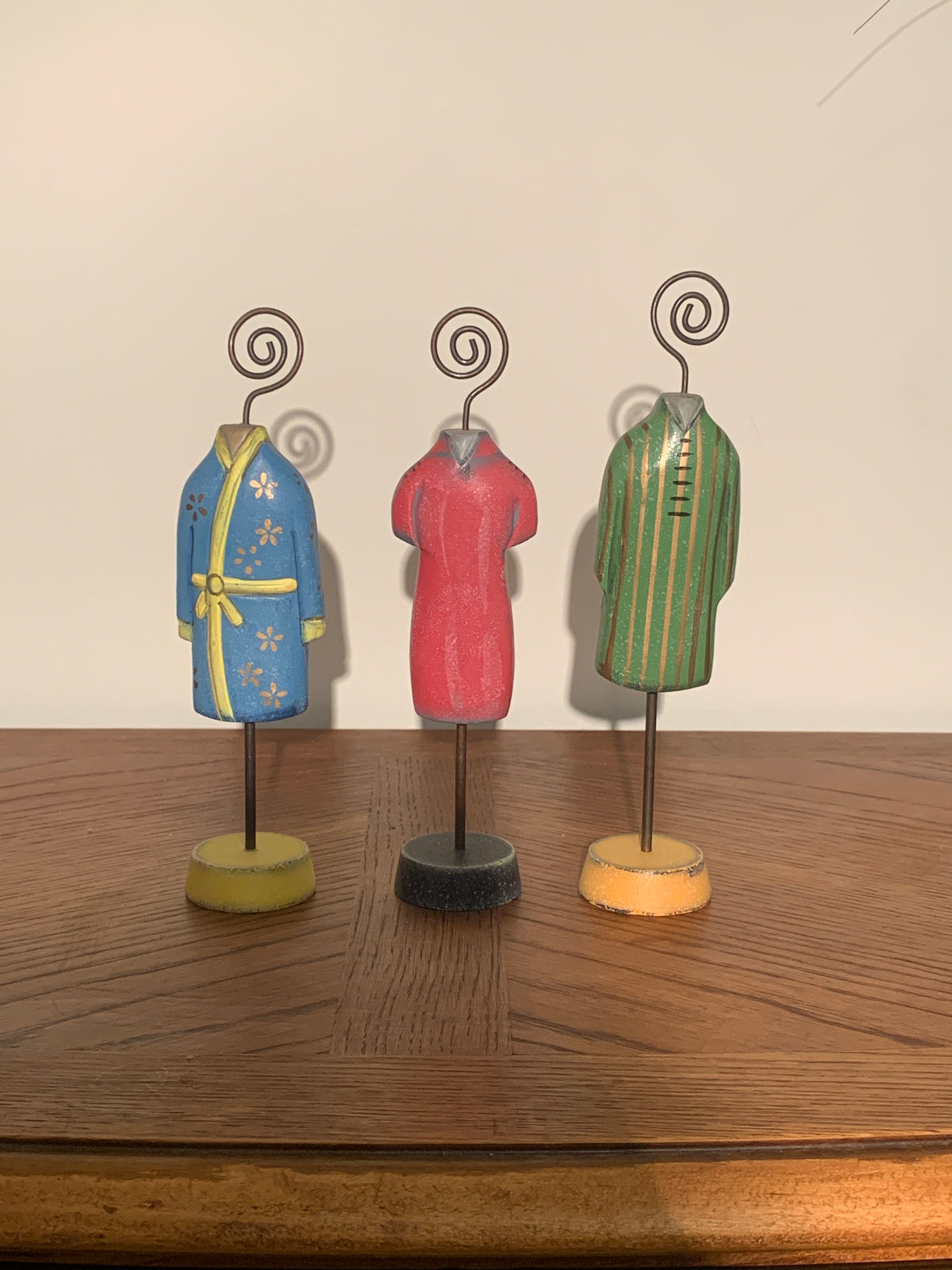 Three small hand-painted miniature clothing mannequins on spiral wire stands, displayed on a wooden table against a plain beige wall. The left figure wears a blue robe with yellow floral patterns and a yellow sash.