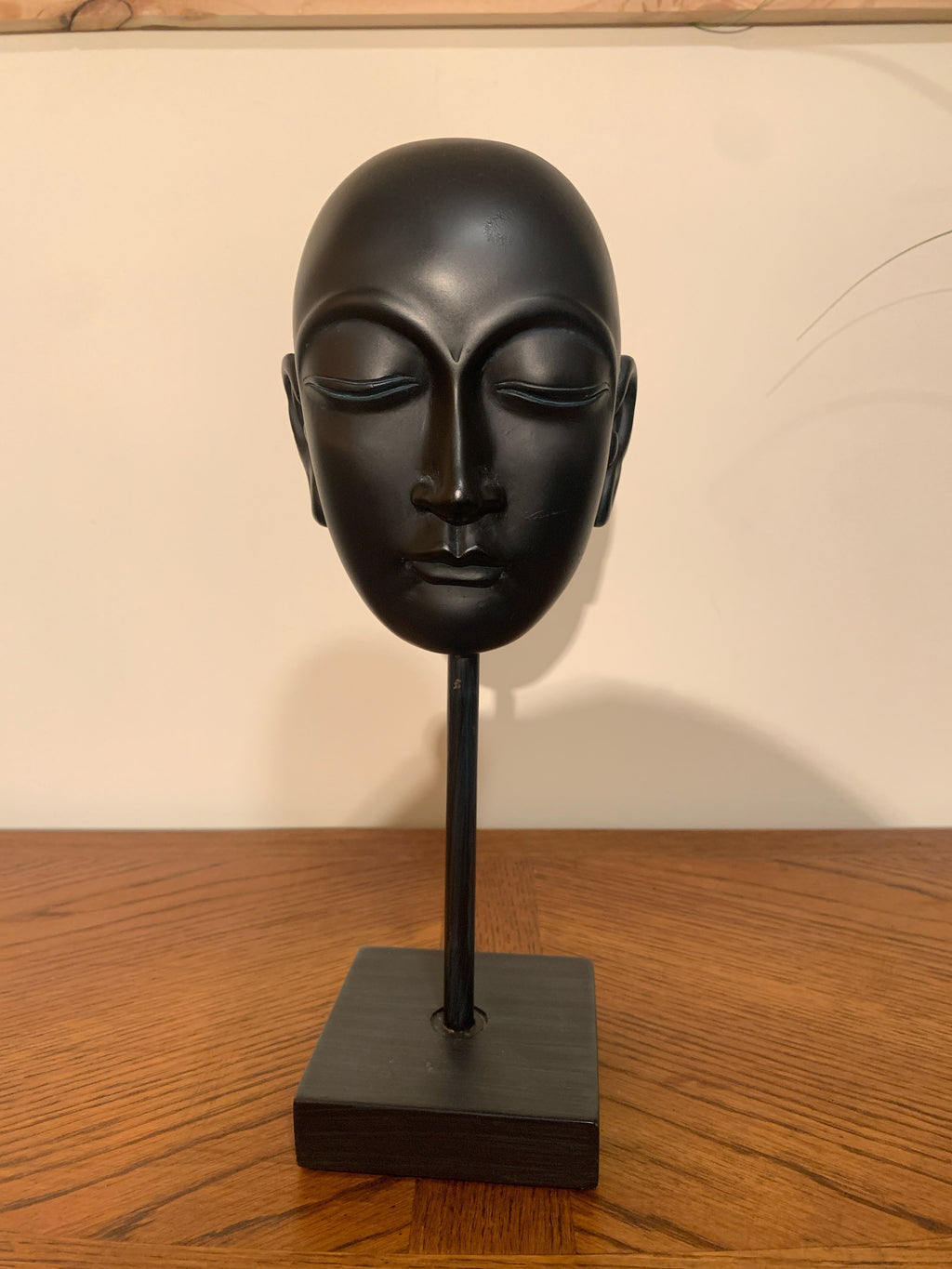 A matte black ceramic or stone Buddha head sculpture displayed on a matching black stand with a square base, set on a wooden tabletop against a plain off-white wall.