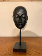 A matte black ceramic or stone Buddha head sculpture displayed on a matching black stand with a square base, set on a wooden tabletop against a plain off-white wall.