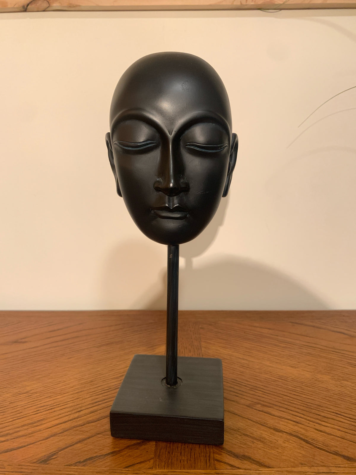 A matte black ceramic or stone Buddha head sculpture displayed on a matching black stand with a square base, set on a wooden tabletop against a plain off-white wall.