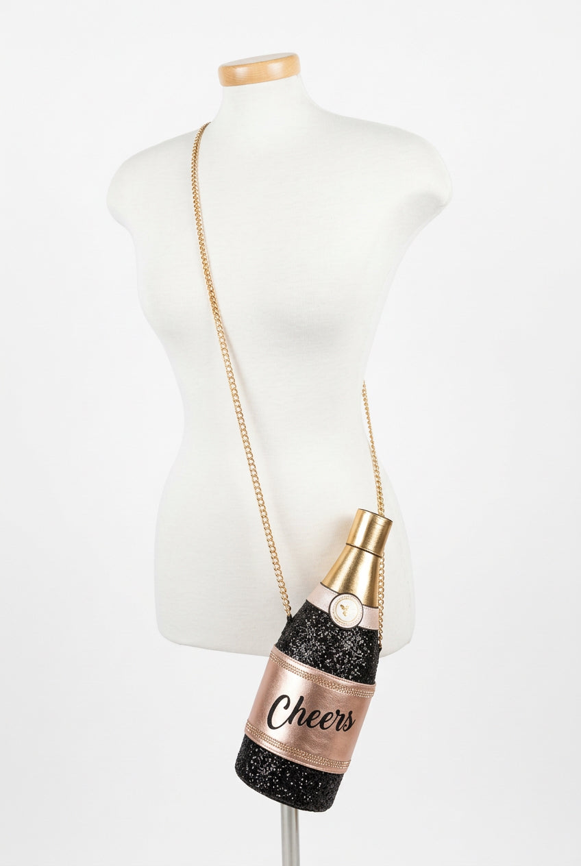A champagne bottle-shaped crossbody bag with a gold tone chain strap, Novelty Aldo Champagne Bottle Purse. The bag features a black glittery body with a pink label that reads 'Cheers' and a gold foil neck.