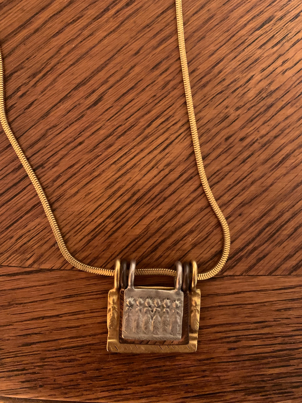 A gold-toned snake chain necklace with a textured padlock pendant, displayed on a dark brown wooden surface.