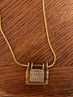 A gold-toned snake chain necklace with a textured padlock pendant, displayed on a dark brown wooden surface.